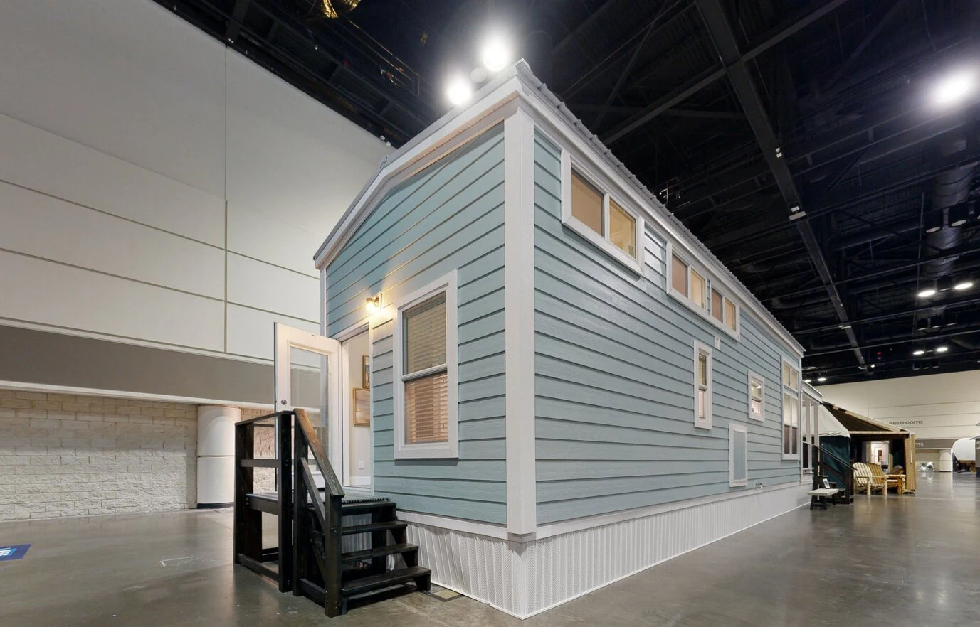 chariot_eagle_ext_1_eg12341a_1703866768787_1378_12 A light blue tiny house with white trim and black steps is on display indoors under large overhead lights.