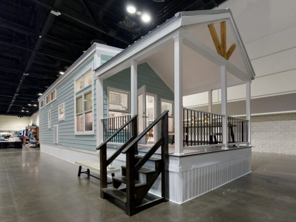 A light blue tiny house with white trim and a covered front porch is displayed indoors at an exhibition hall.