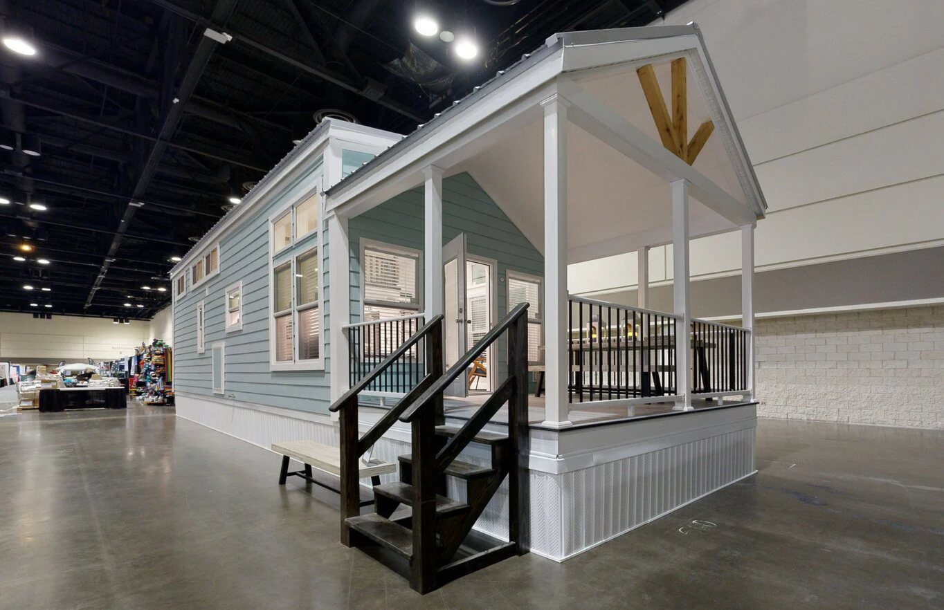 chariot_eagle_ext_2_eg12341a_1703866765619_1364_12 A light blue tiny house with white trim and a covered front porch is displayed indoors at an exhibition hall.