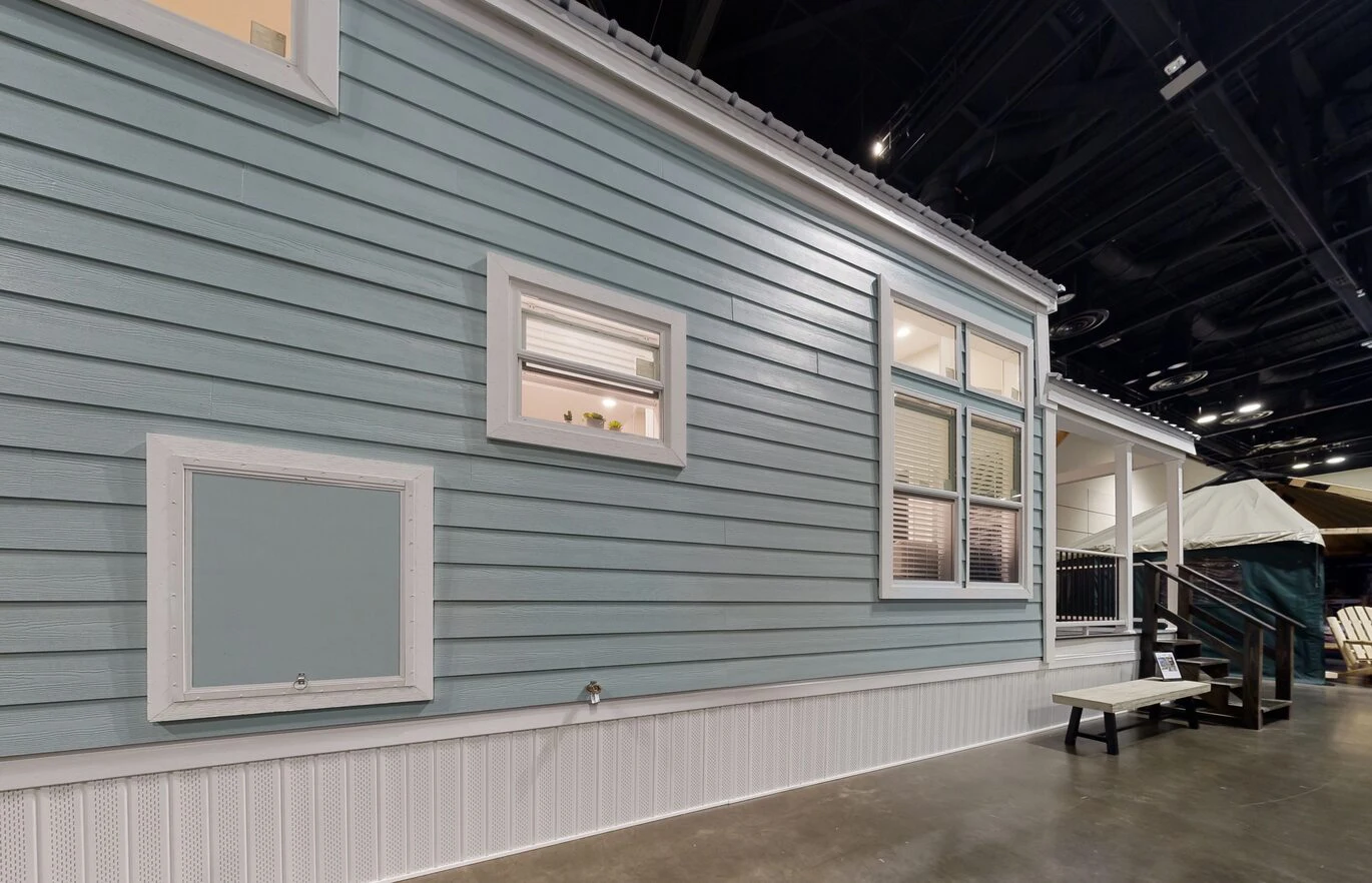 chariot_eagle_ext_3_eg12341a_1703866763253_1370_12 A light blue house with white trim and windows is displayed indoors on a concrete floor with a bench and porch visible on the right side.