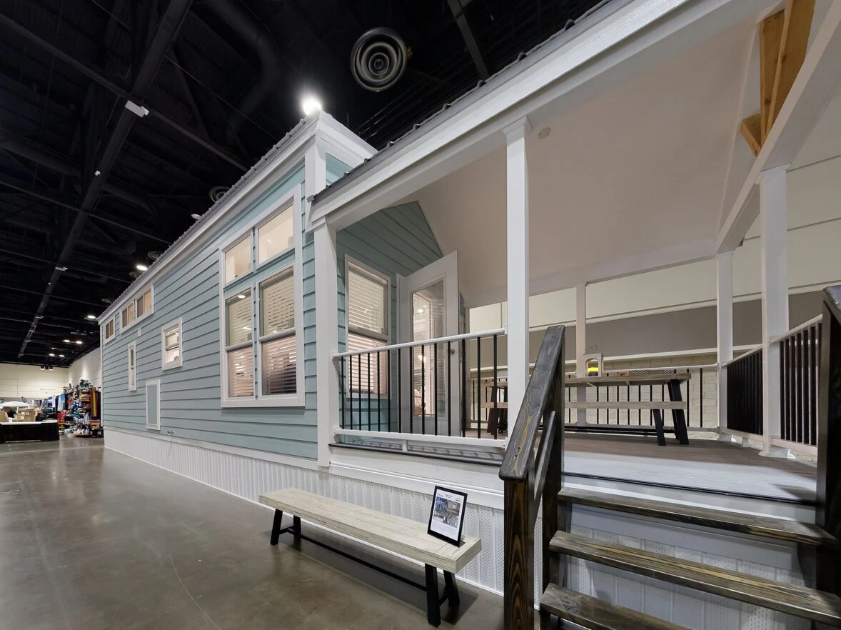 chariot_eagle_ext_4_eg12341a_1703866760098_1178_12 A light blue tiny house with white trim is displayed indoors, featuring a covered porch with stairs, railing, and a bench in front.