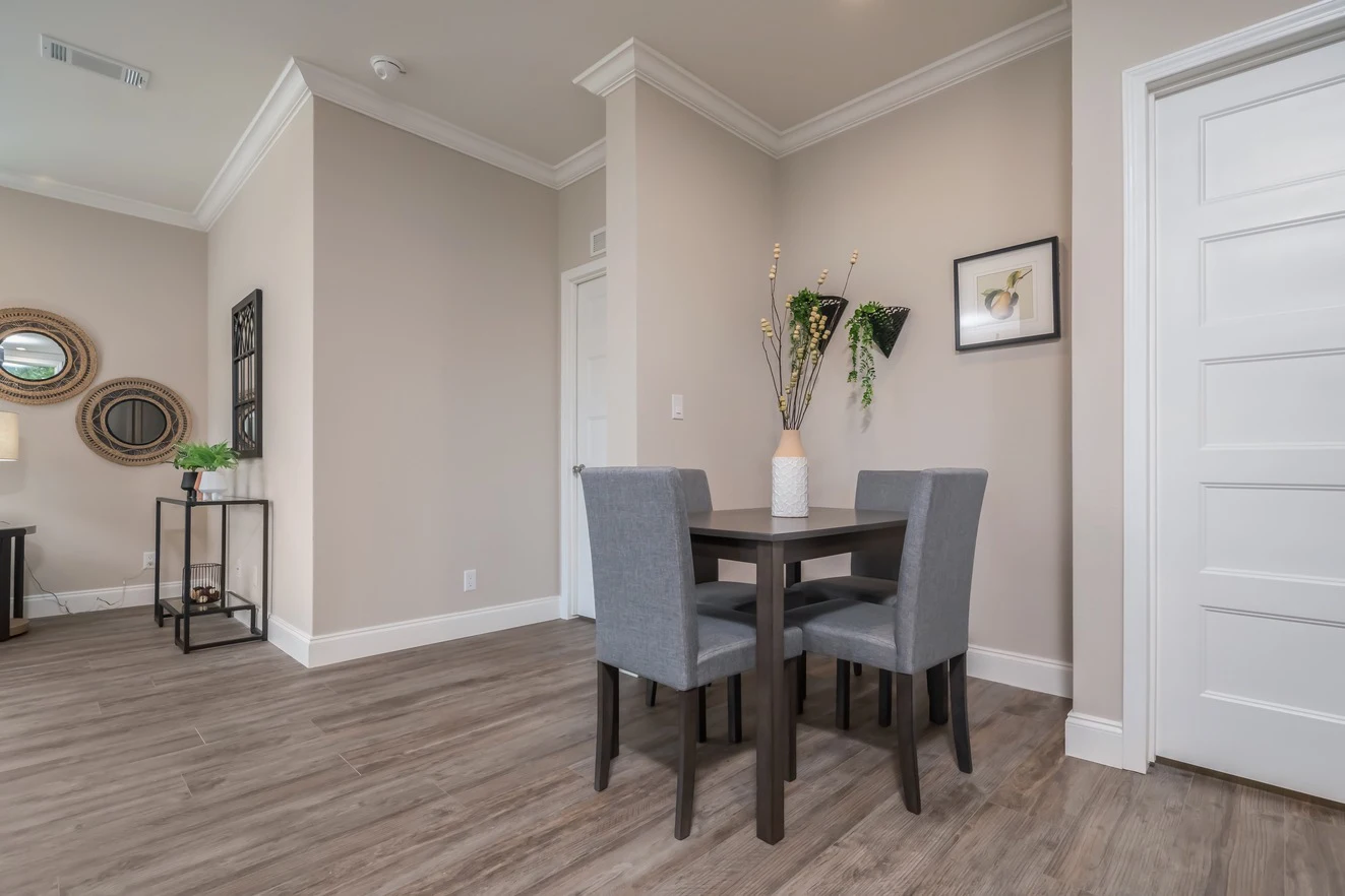 A small dining area with a square dark wood table, four gray upholstered chairs, a vase with decorative branches, wall art, and light wood flooring in a modern apartment.