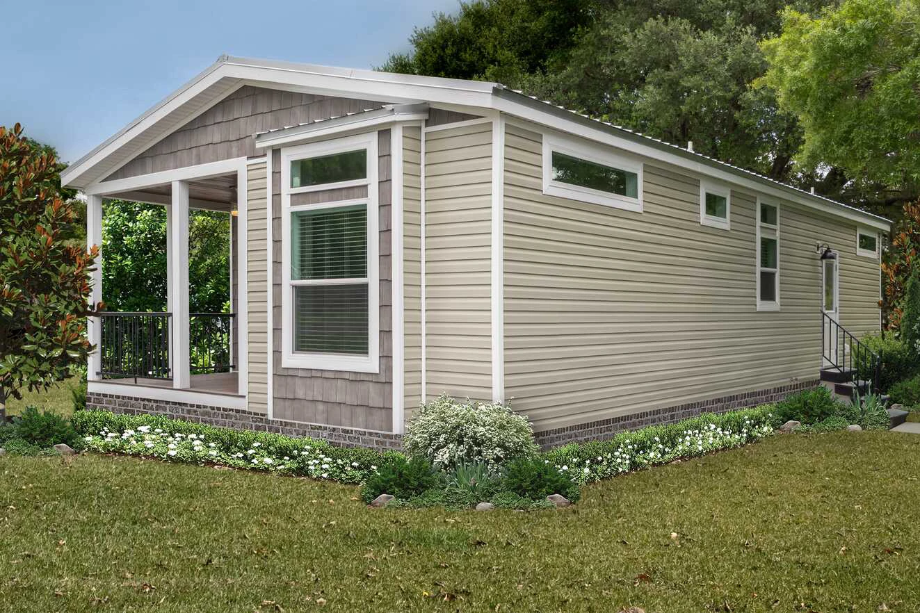 A small beige manufactured home with a covered front porch, surrounded by green grass, landscaping, and trees in the background.