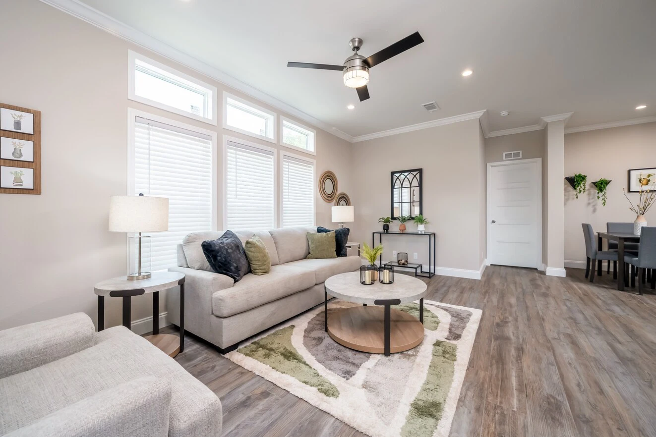 Modern living room with light-colored furniture, patterned rug, round coffee table, wall art, ceiling fan, and large windows; dining area visible in the background.