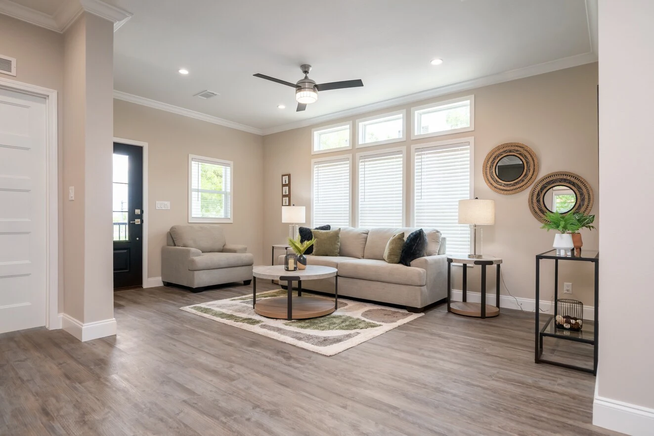 Modern living room with wood flooring, neutral walls, two beige sofas, a round coffee table, lamps, wall art, and large windows letting in natural light.