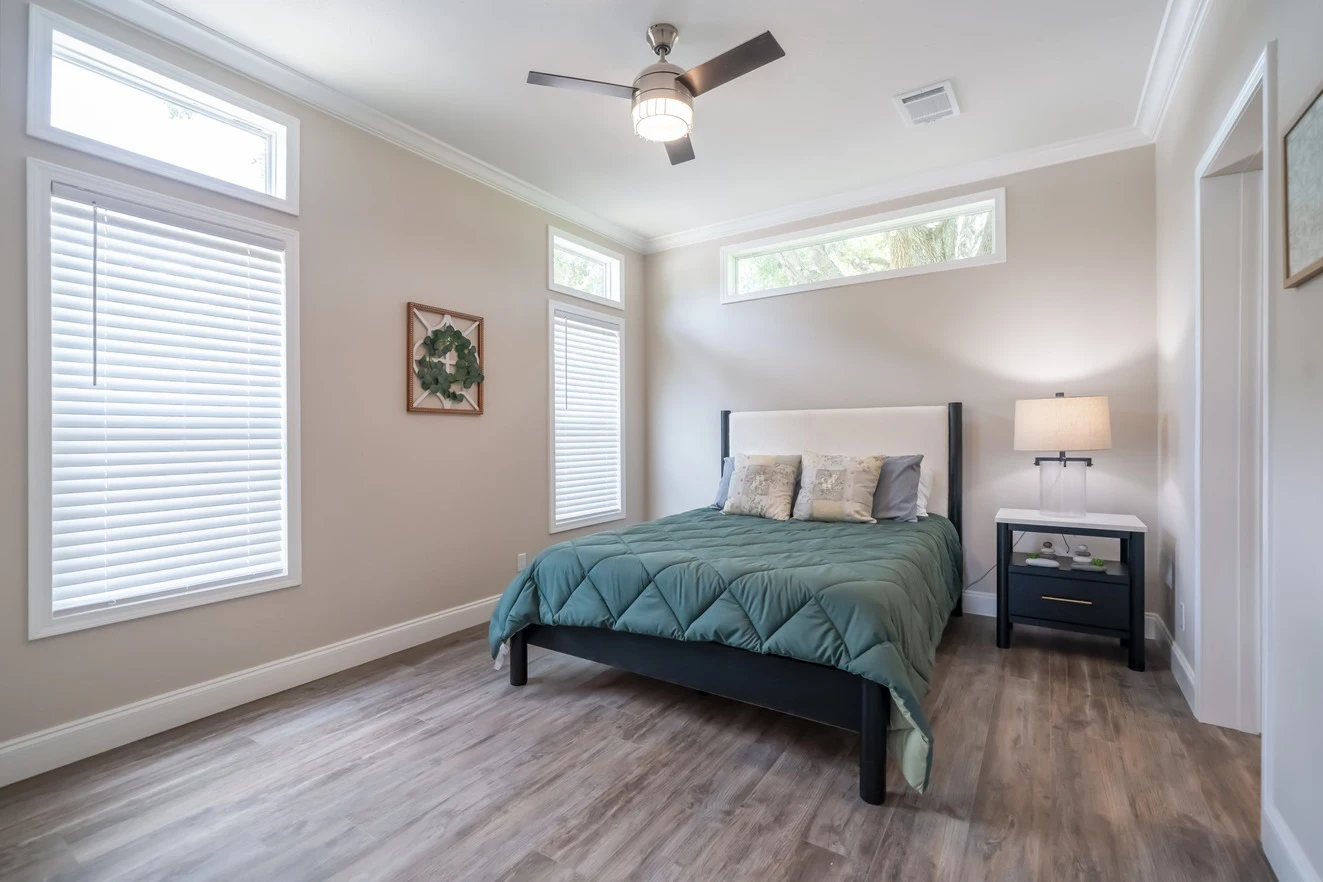 A modern bedroom with a bed, green comforter, two pillows, nightstand with a lamp, three tall windows, ceiling fan, and light wood flooring.