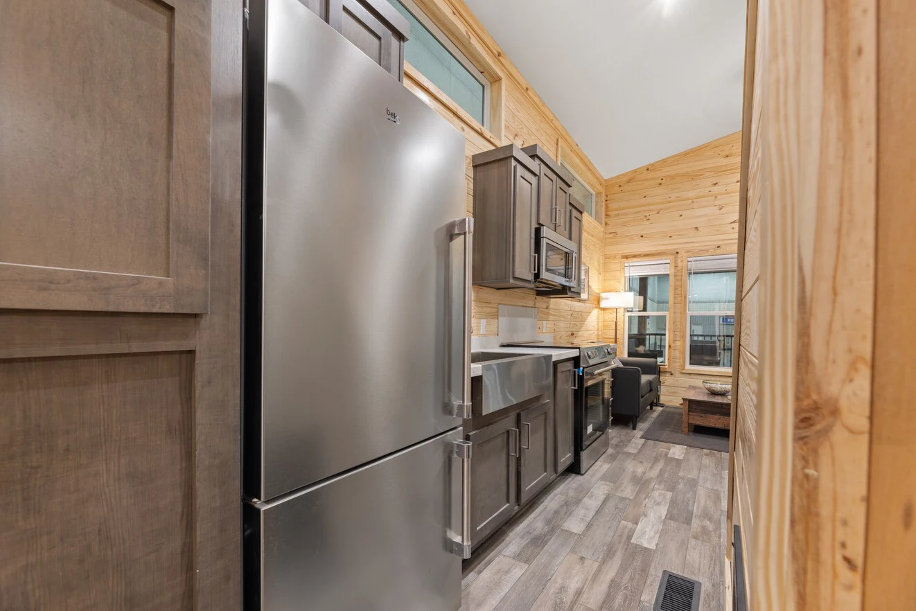 Modern galley kitchen with stainless steel refrigerator, dark cabinets, farmhouse sink, microwave, and stove, set in a wood-paneled tiny house with plank flooring.