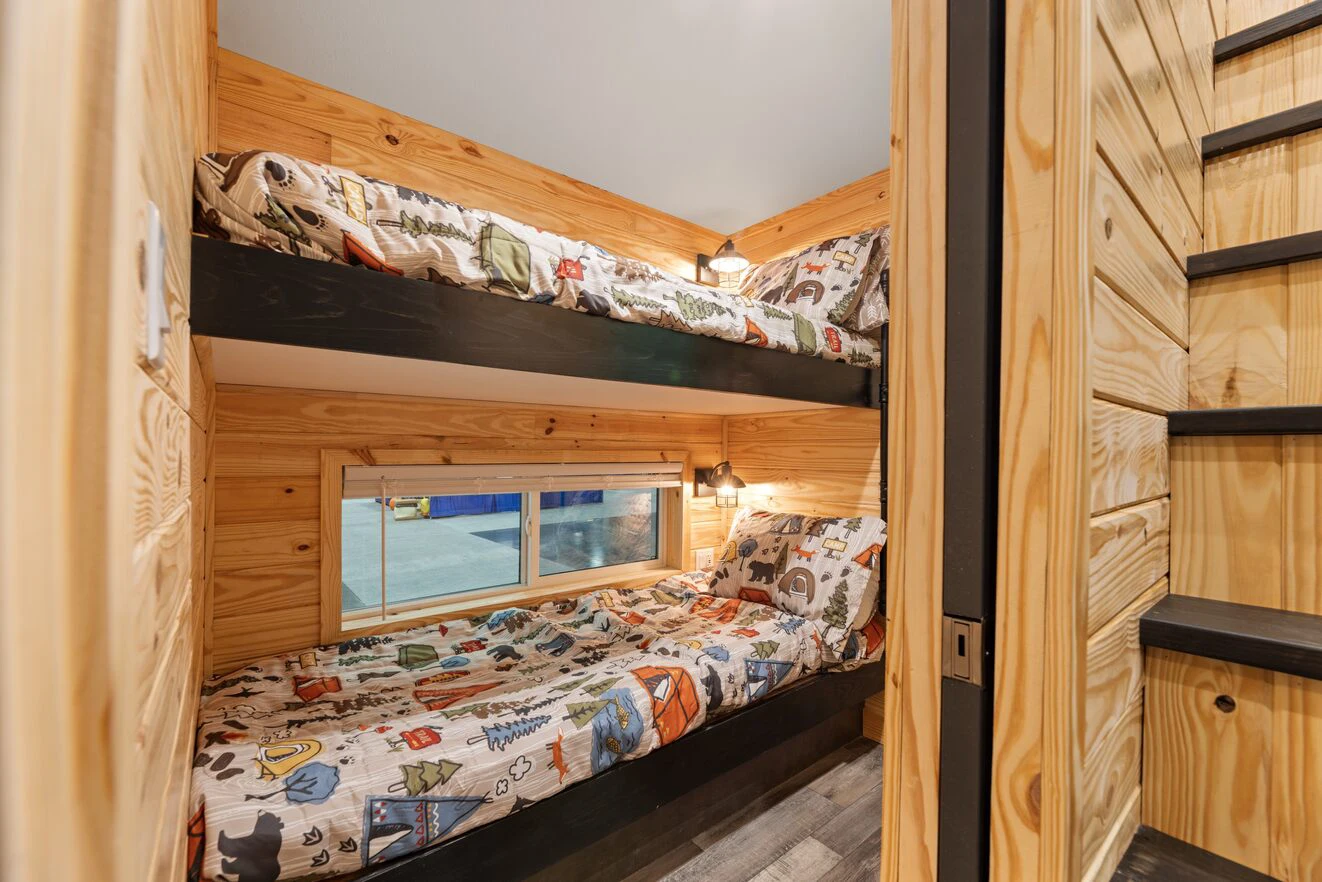 Two wooden bunk beds with patterned bedding are built into a small room with pine walls, a window, and black stairs leading to an upper level.