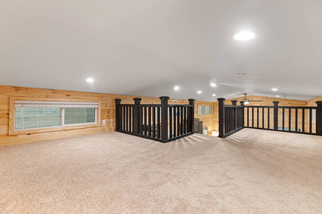 Carpeted loft area with wood-paneled walls, black railing, ceiling lights, and a window on the left side.
