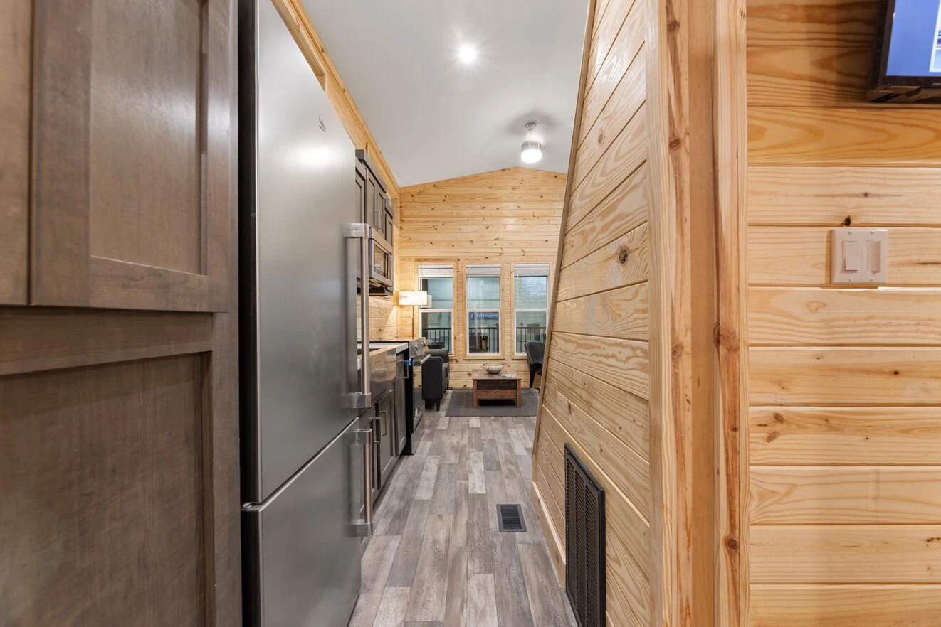 Narrow view of a modern tiny house interior featuring wooden walls, grey cabinetry, stainless steel refrigerator, and a small living area with windows and a sofa at the far end.