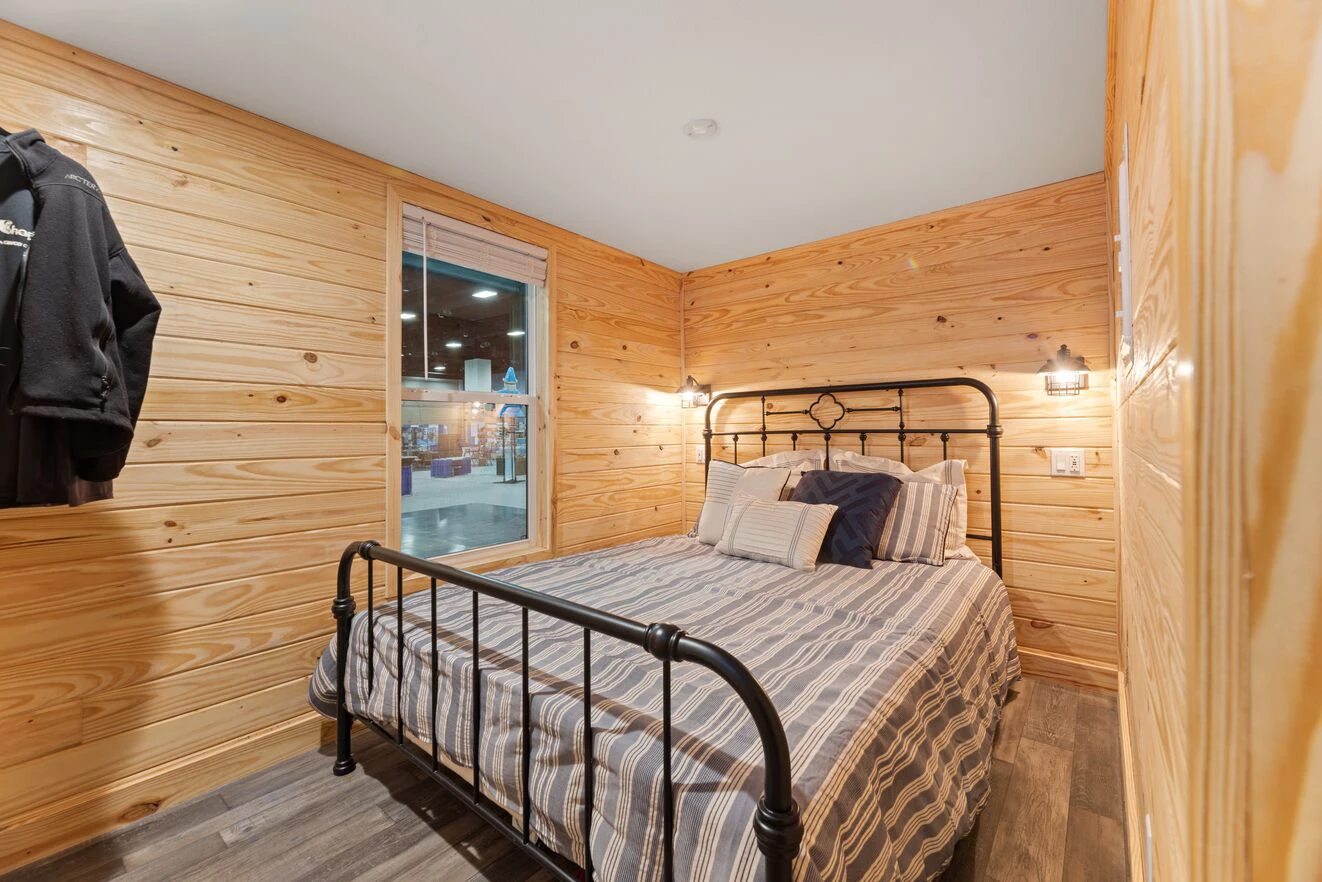 A small bedroom with wood-paneled walls, a metal bed frame, striped bedding, two wall-mounted lamps, and a window showing an indoor space outside.