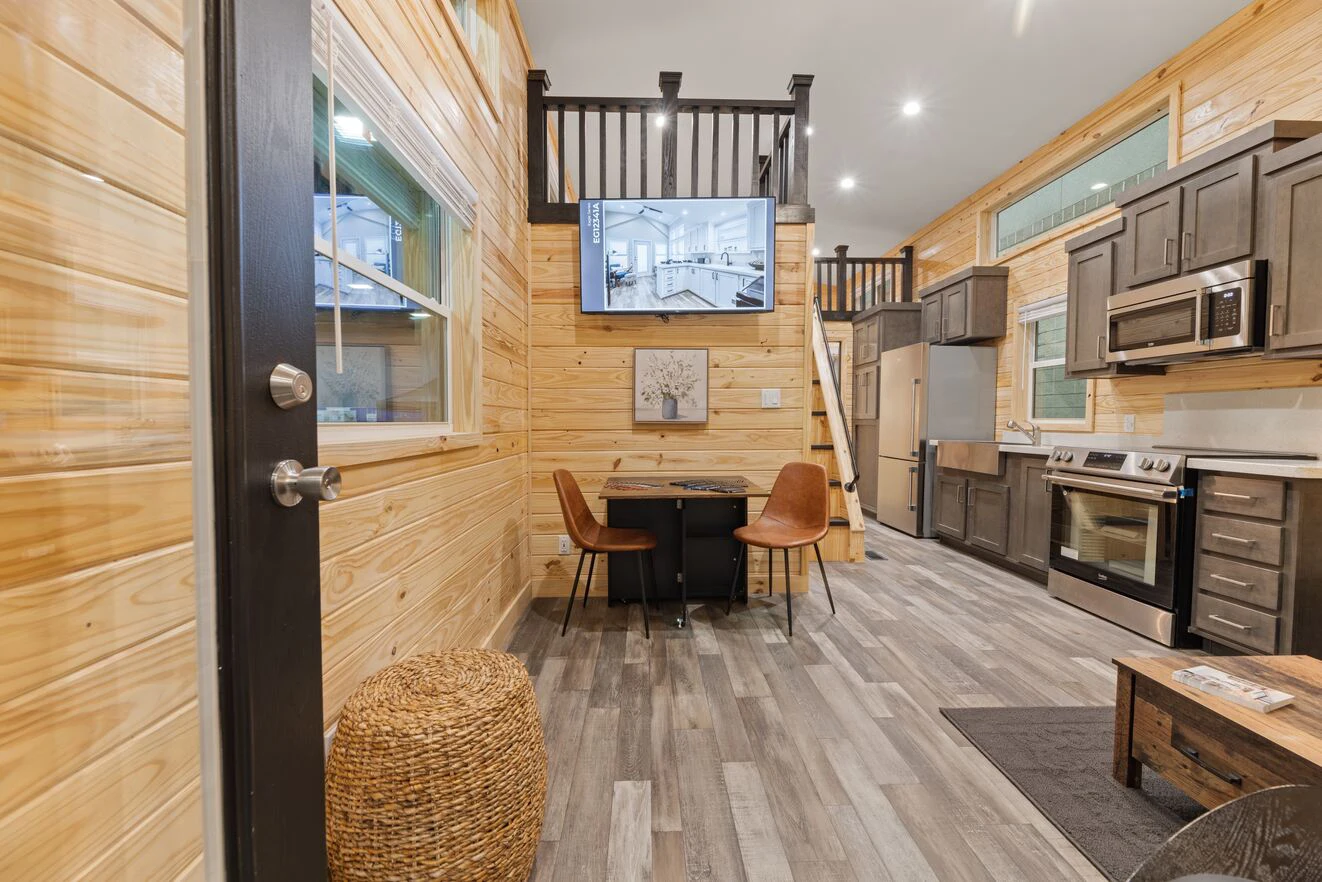 Modern tiny house interior with wood-paneled walls, a kitchen, dining area with two chairs, loft space, and a flat-screen TV mounted above the table.