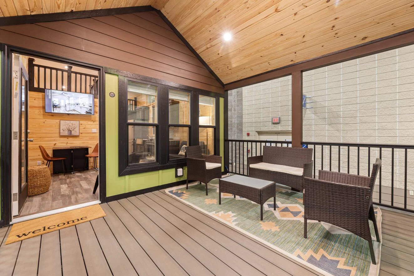 Covered patio with wicker seating, a small table, and a rug. Doorway leads to an indoor office space with a TV and wood-paneled walls. "Welcome" mat at the entrance.