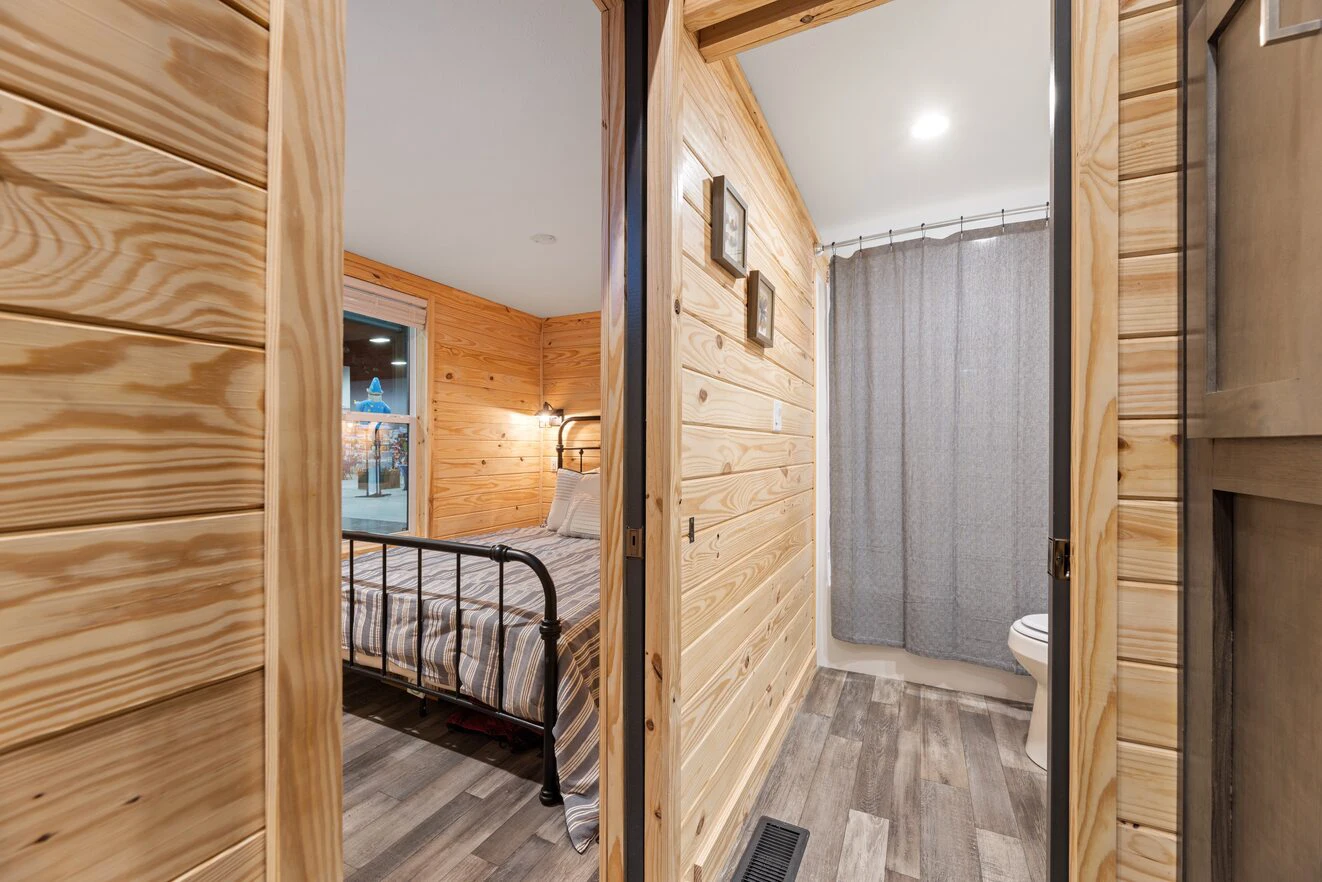 View of a small wooden cabin interior showing a bedroom with a metal bed frame on the left and a bathroom with a gray shower curtain and toilet on the right.