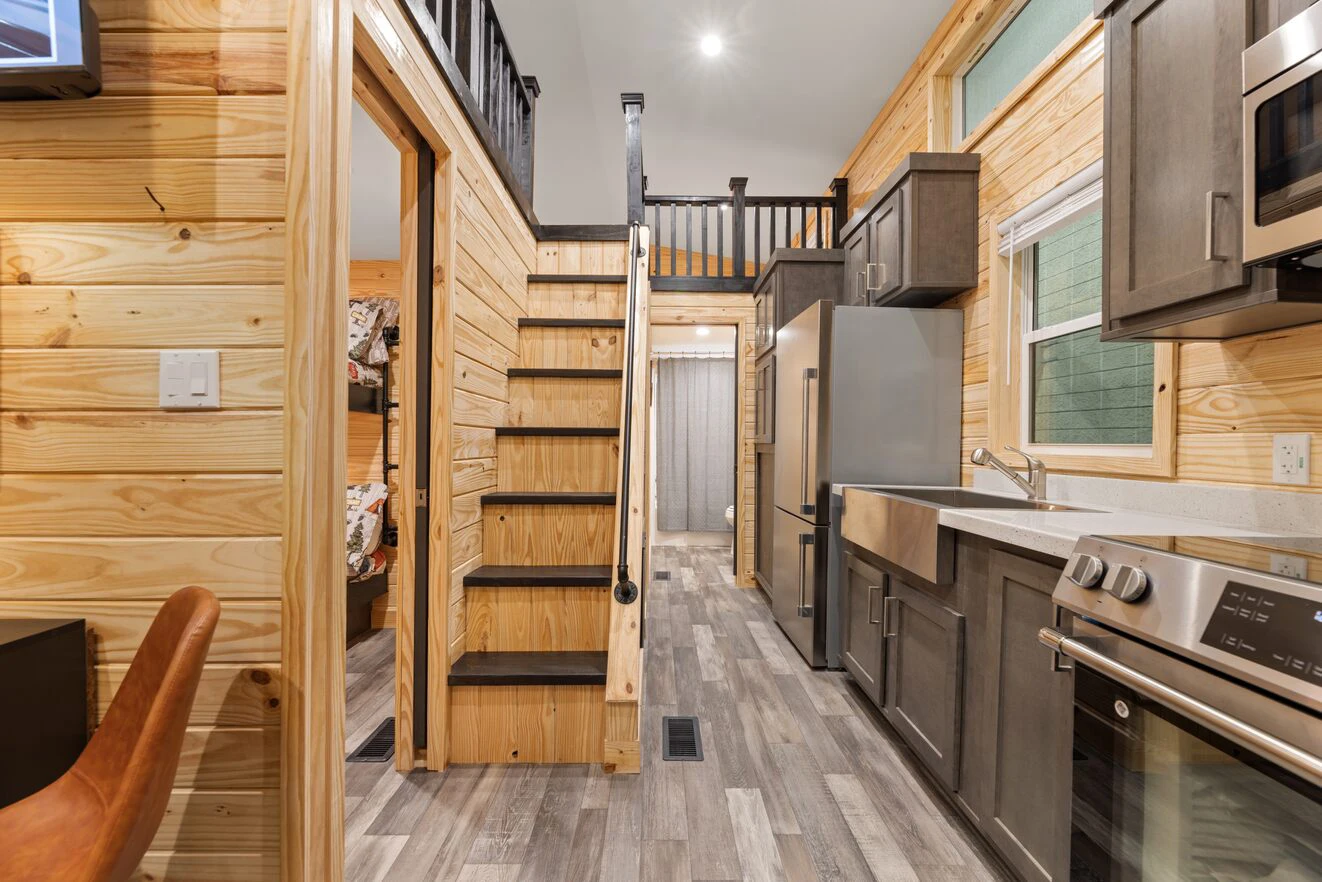 Compact modern kitchen with wooden walls, grey cabinets, stainless steel appliances, and a staircase leading to a loft area; bunk beds visible in a side room.