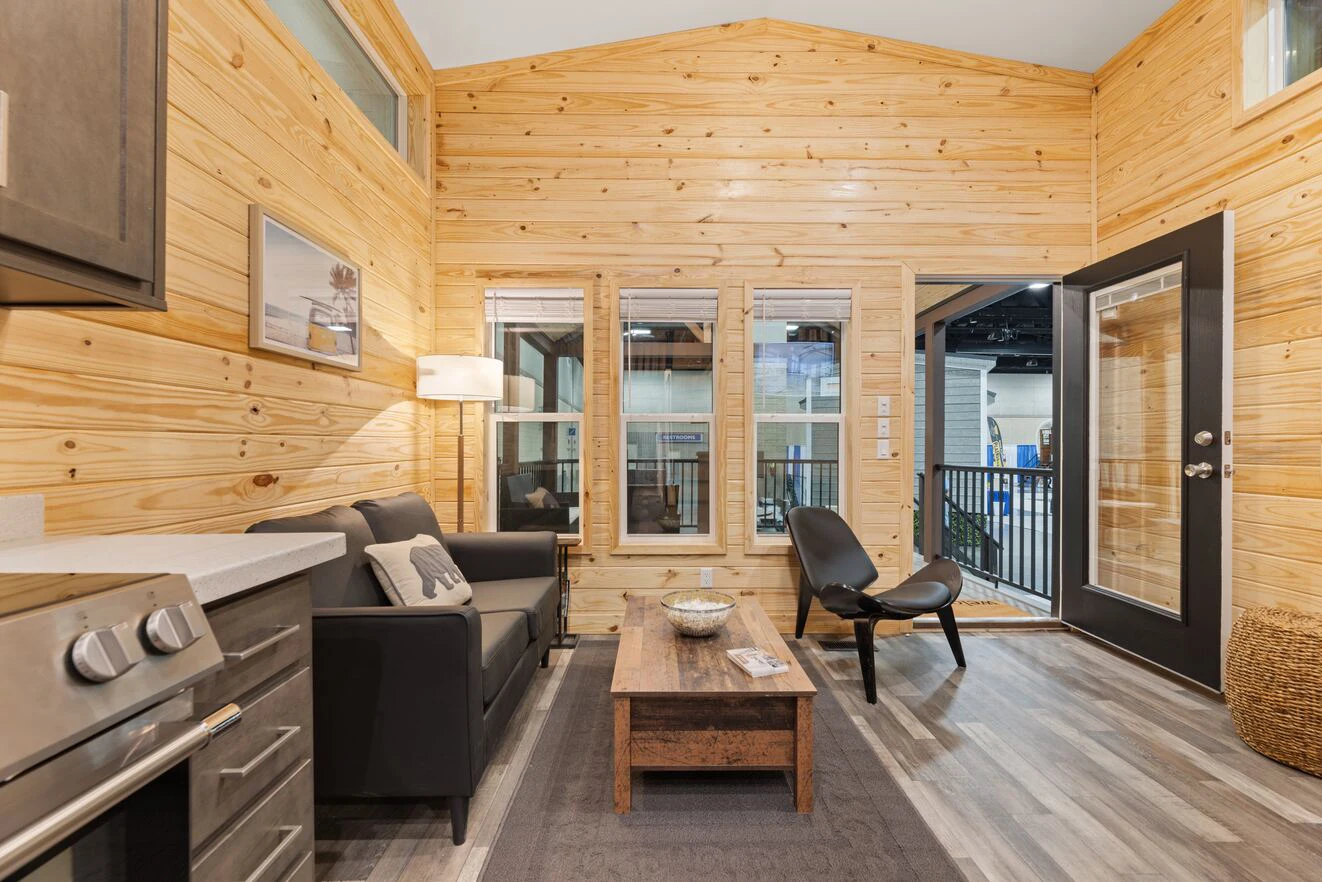 A small living area with wood-paneled walls, a black sofa, a wooden coffee table, a black chair, and an open glass door leading to a deck.