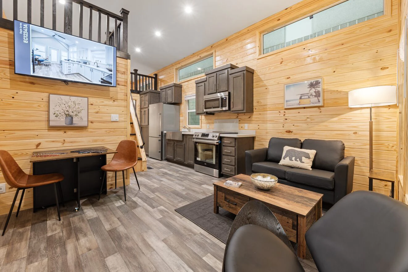 A modern tiny house interior with wooden walls, a kitchen, dining table, sofa, coffee table, TV, and loft area visible above.