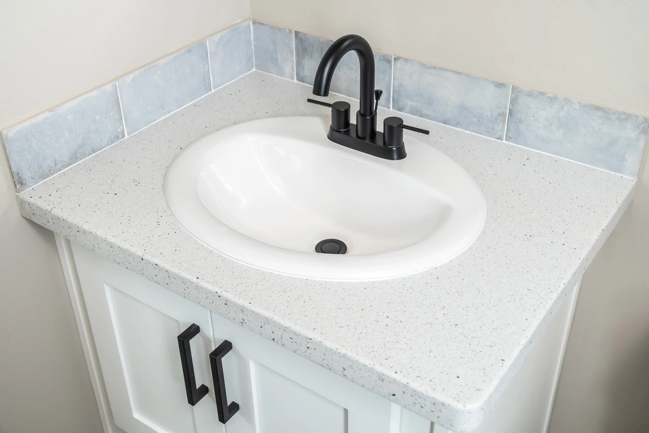 White bathroom sink with black faucet and handles, set in a speckled countertop with blue-gray tile backsplash and white vanity cabinet below.