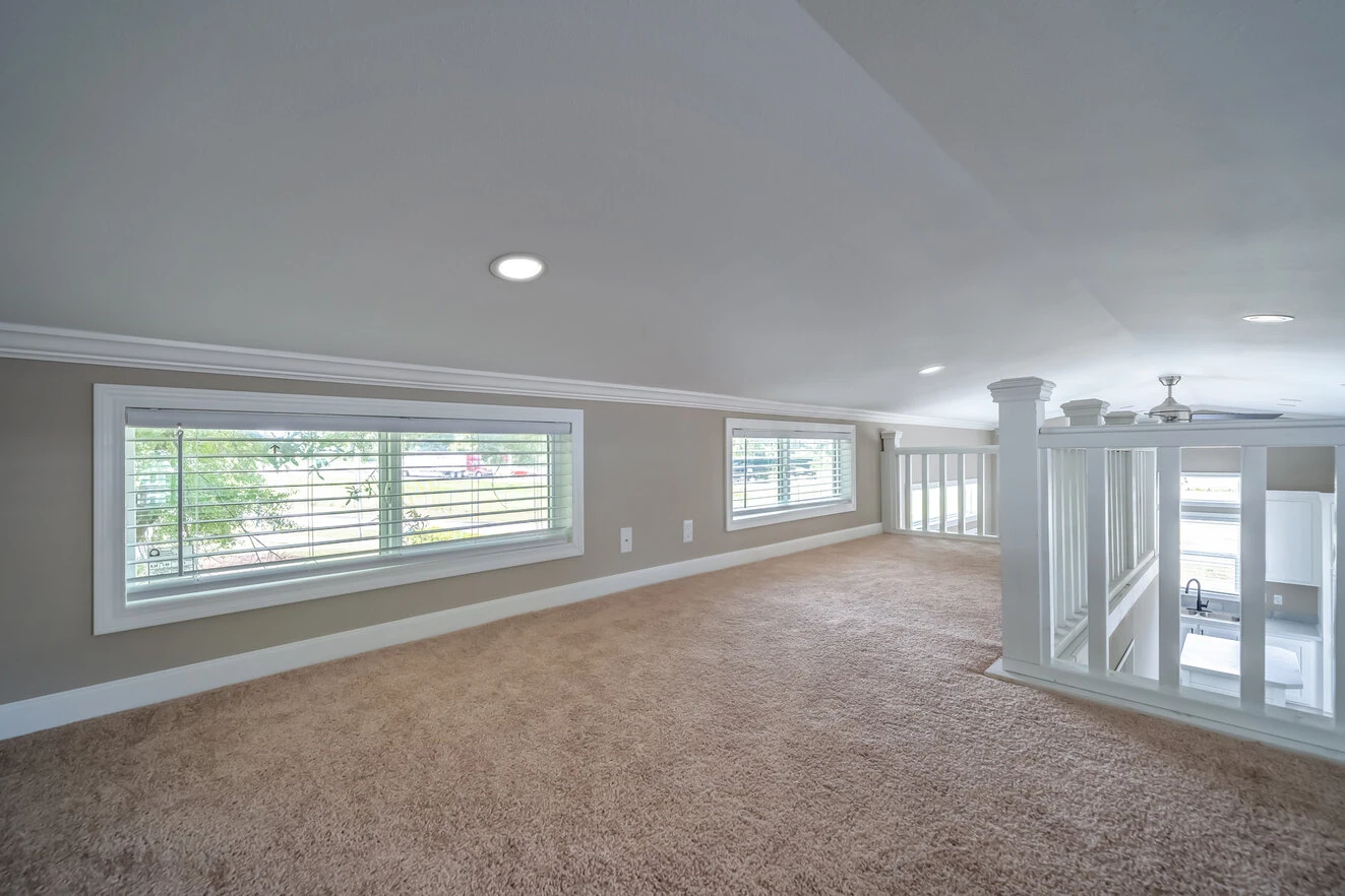 A carpeted loft area with three rectangular windows, beige walls, and white trim, overlooking a lower level with a ceiling fan and staircase.