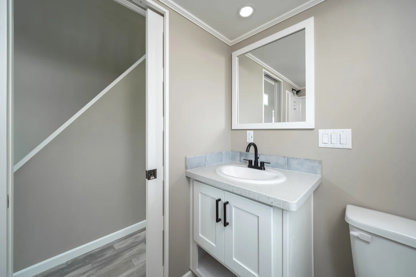 A modern bathroom with a white vanity, black faucet, wall mirror, and toilet. The door is open, revealing a hallway with gray flooring and beige walls.