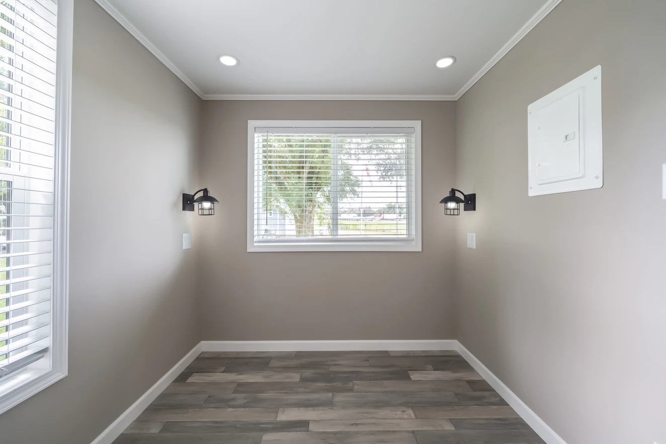 A small empty room with beige walls, a window with blinds, two wall-mounted lights, wood-patterned flooring, and a white electrical panel.