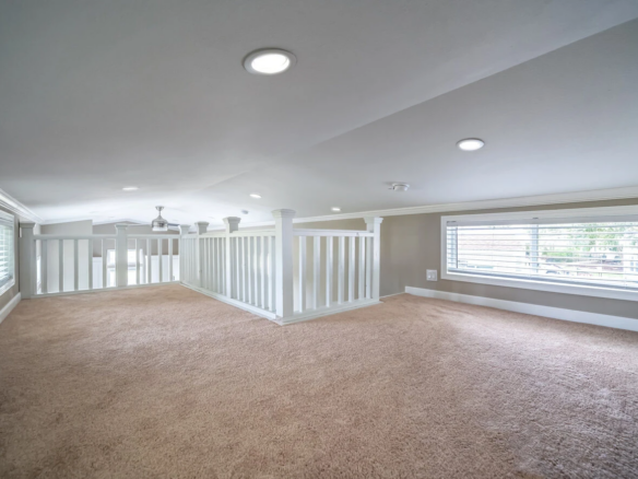 A spacious, carpeted loft area with white railings, recessed ceiling lights, and windows with shutters on both sides, providing ample natural light.
