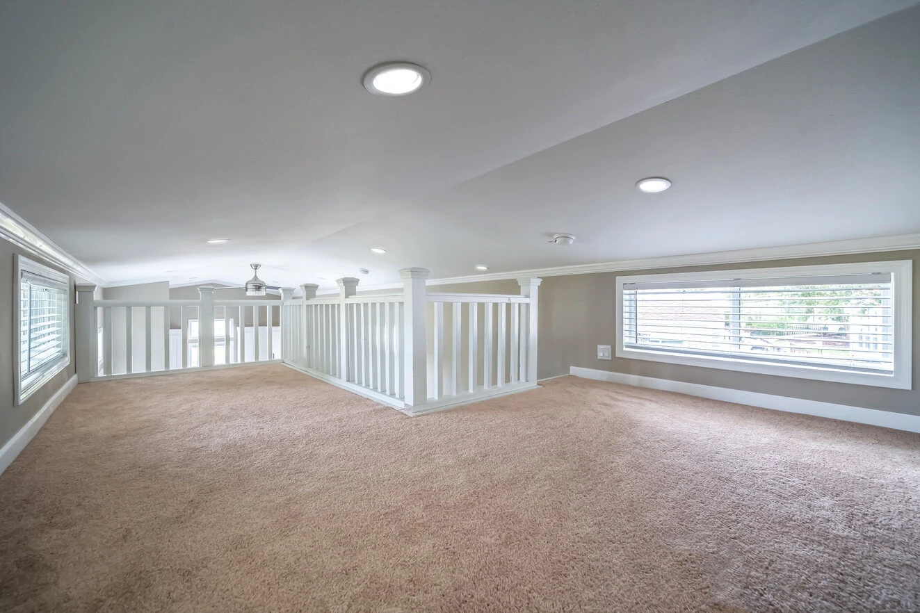 A spacious, carpeted loft area with white railings, recessed ceiling lights, and windows with shutters on both sides, providing ample natural light.