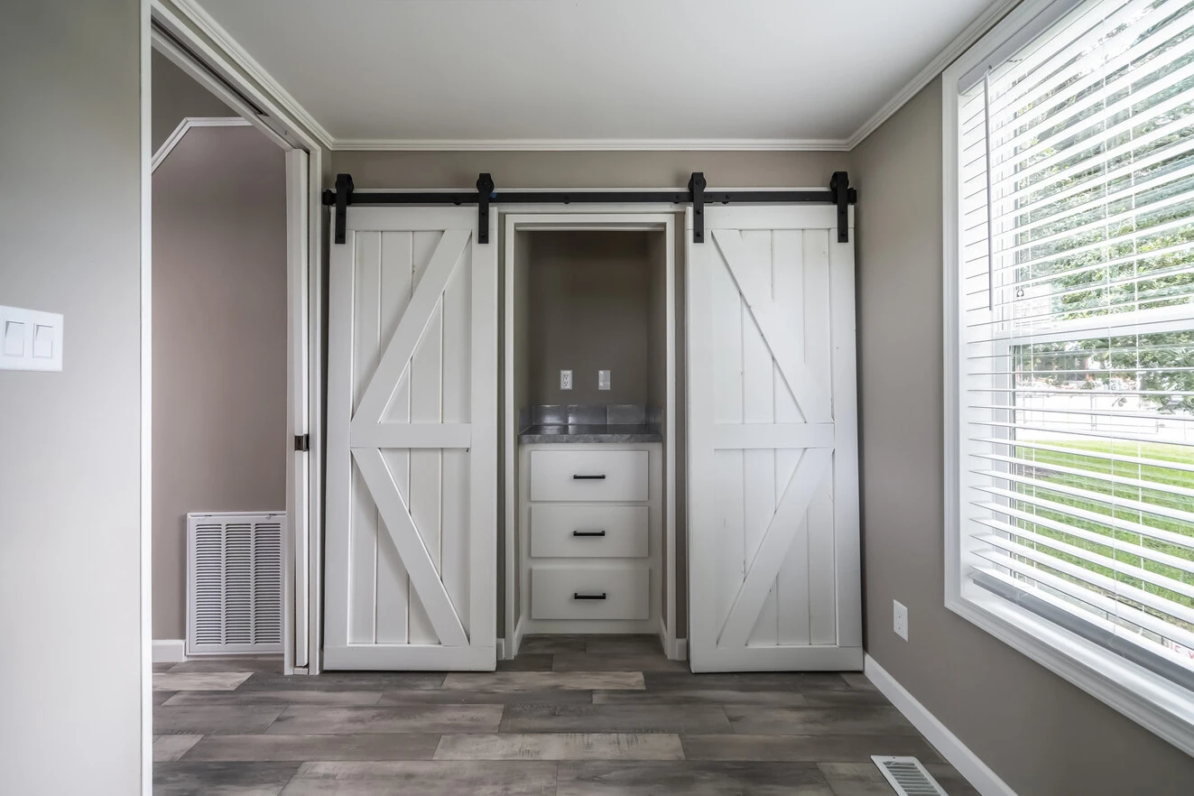 A small closet with white sliding barn doors, three drawers, and a countertop is in a room with gray walls, wood-look flooring, and a large window with blinds.