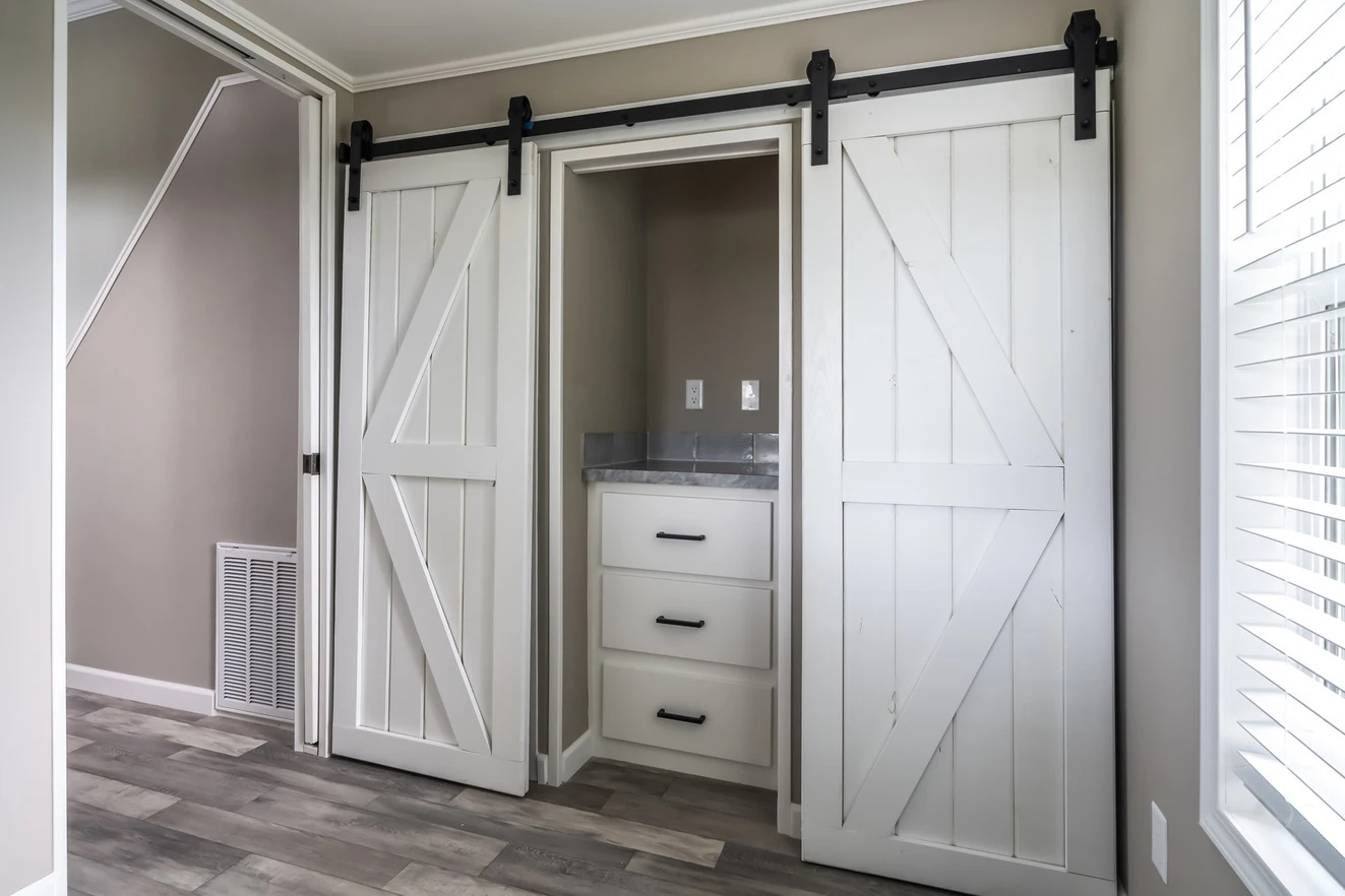 Small alcove with a sink and drawers is enclosed by white sliding barn doors inside a light gray room with wood flooring.