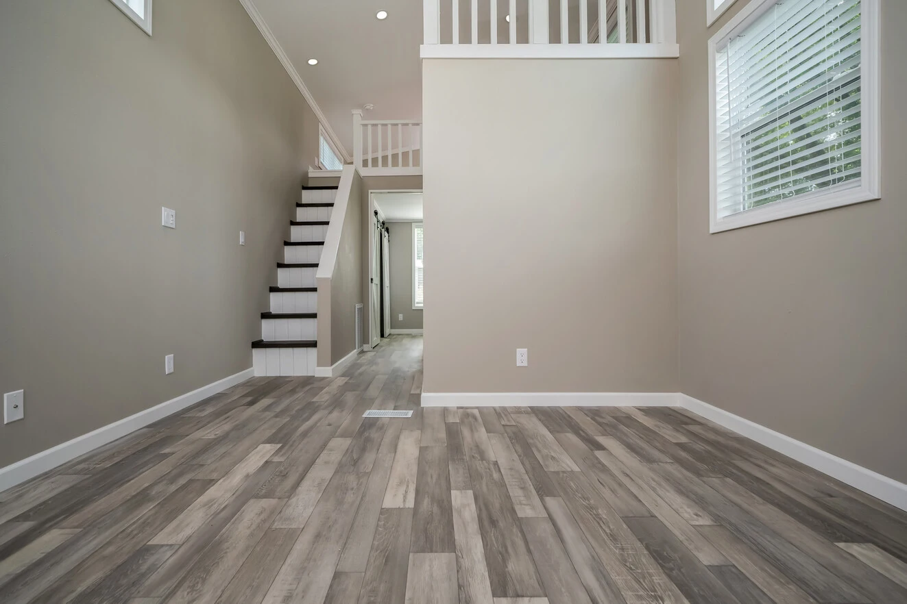 Empty room with light wood flooring, beige walls, white trim, staircase with dark steps, and multiple windows with blinds.