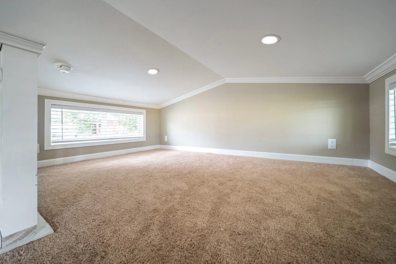 Low-ceilinged room with beige carpet, light taupe walls, recessed lighting, and a wide window with blinds.