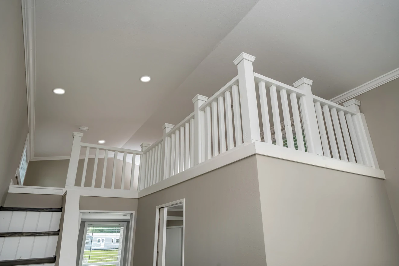 Second-story loft area with white railings, beige walls, recessed ceiling lights, and a stairway leading up from the lower floor.