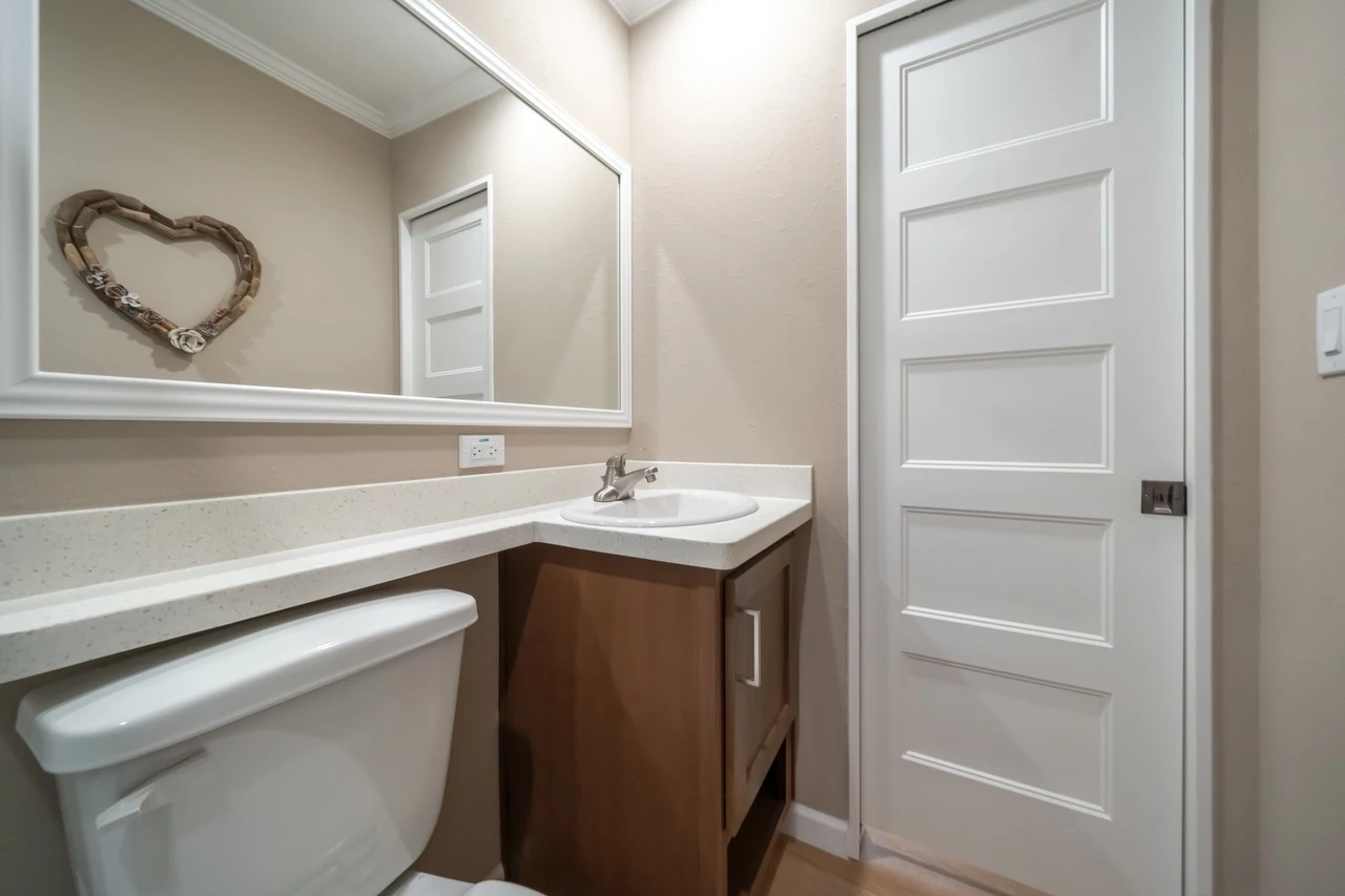 Small bathroom with a white toilet, single sink vanity, large framed mirror, beige walls, and a white paneled door. A heart-shaped decoration hangs on the wall.