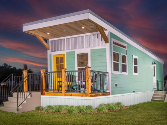 A modern tiny house with mint green siding, a yellow door, a small porch with wooden railings, and exterior lights, set on a grassy lawn at dusk.