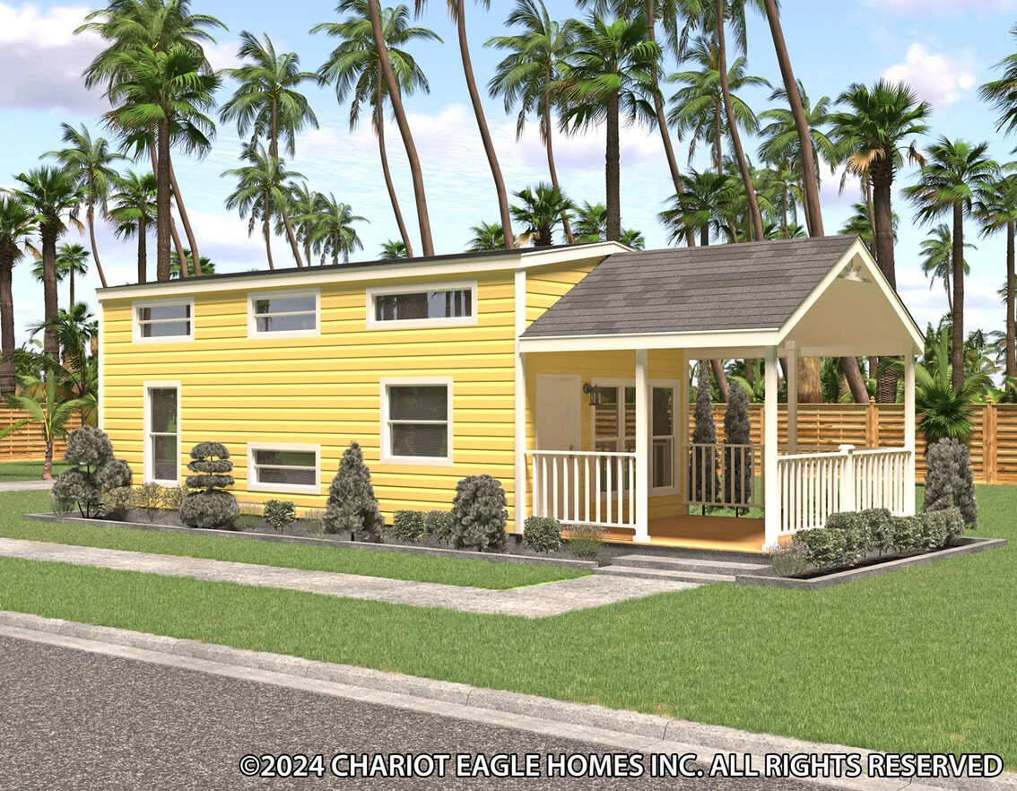 A yellow tiny house with white trim, a front porch, small shrubs, and palm trees in the background. The home sits on a manicured lawn beside a paved road.