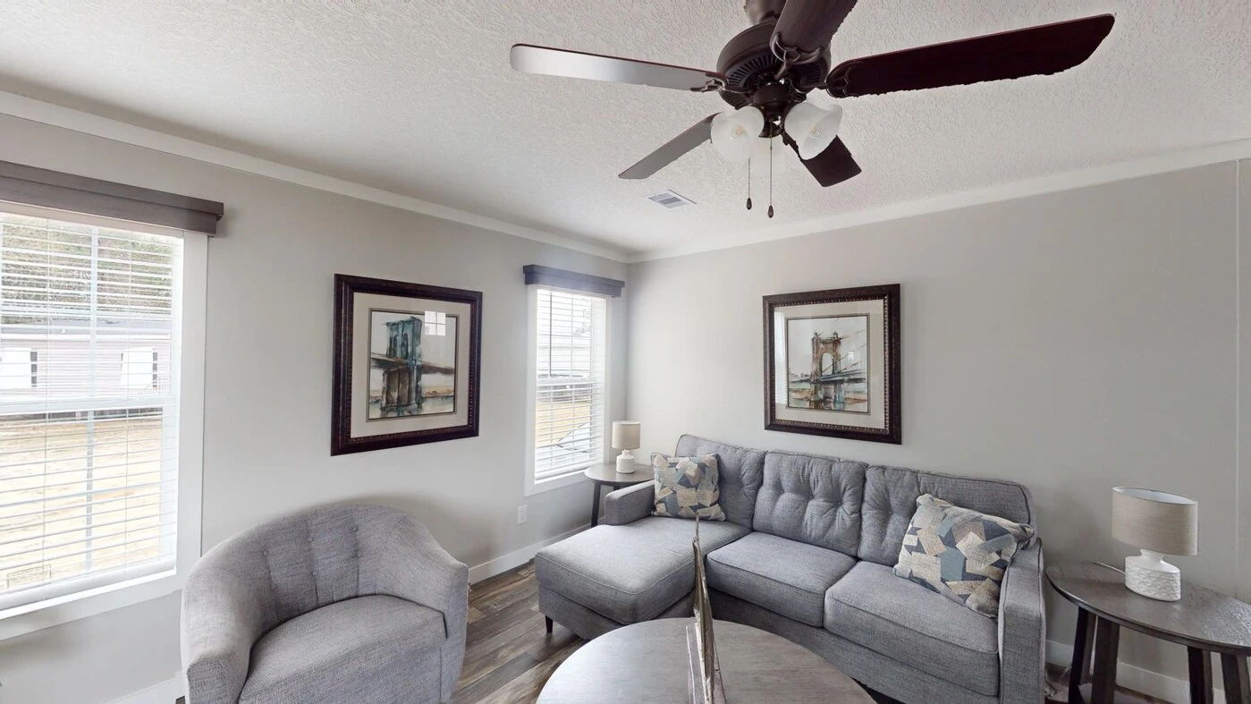 A modern living room with a gray sectional sofa, round armchair, coffee table, ceiling fan, two framed pictures, and large windows with white blinds.