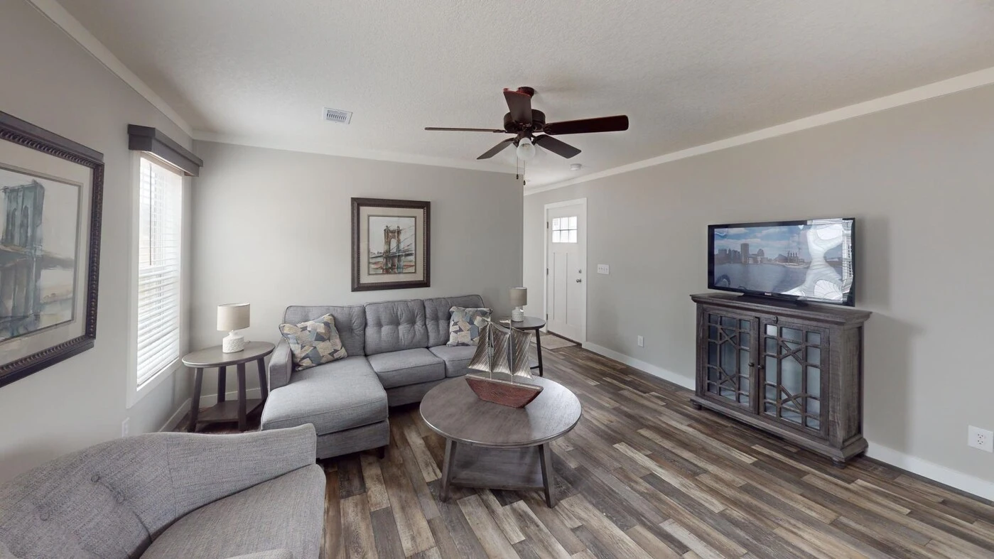A modern living room with gray furniture, wood flooring, a ceiling fan, wall art, and a TV on a cabinet against a light gray wall.