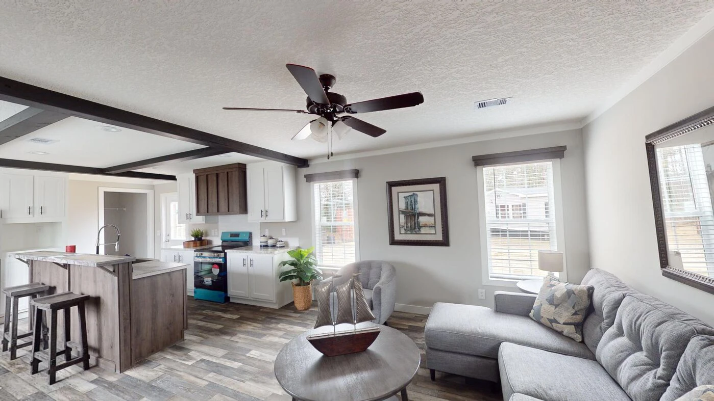 A modern open-plan living room and kitchen with a gray sectional sofa, round coffee table, ceiling fan, and white cabinets. Large windows let in natural light.