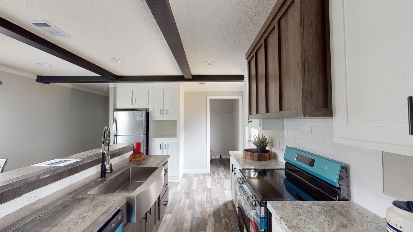 Modern kitchen with stainless steel appliances, double sink, wood cabinets, white backsplash, and exposed ceiling beams. Light wood flooring and a small plant on the counter.