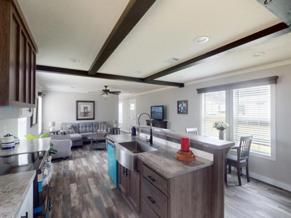 Open-concept kitchen with wood cabinets and island, stainless steel appliances, and farmhouse sink overlooking a living room with a gray sectional sofa and large windows.