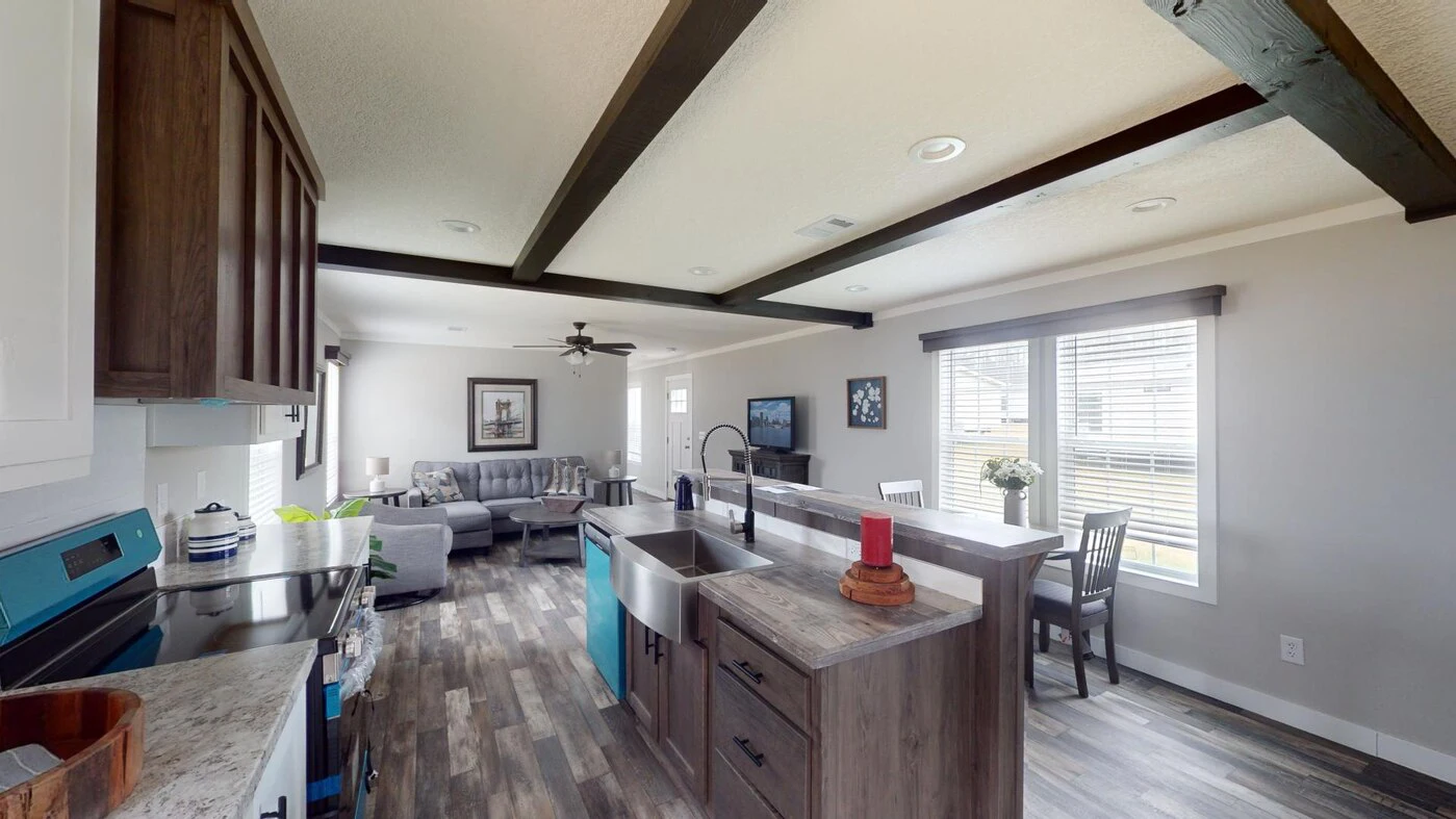 Open-concept kitchen with wood cabinets and island, stainless steel appliances, and farmhouse sink overlooking a living room with a gray sectional sofa and large windows.