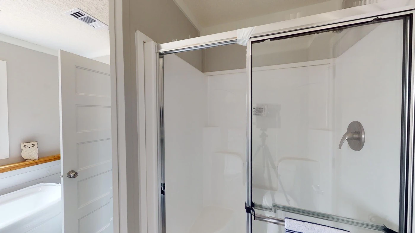 A bathroom with a glass shower enclosure, a silver showerhead, a white towel on the door, and an open door revealing part of a bathtub with an owl decoration.