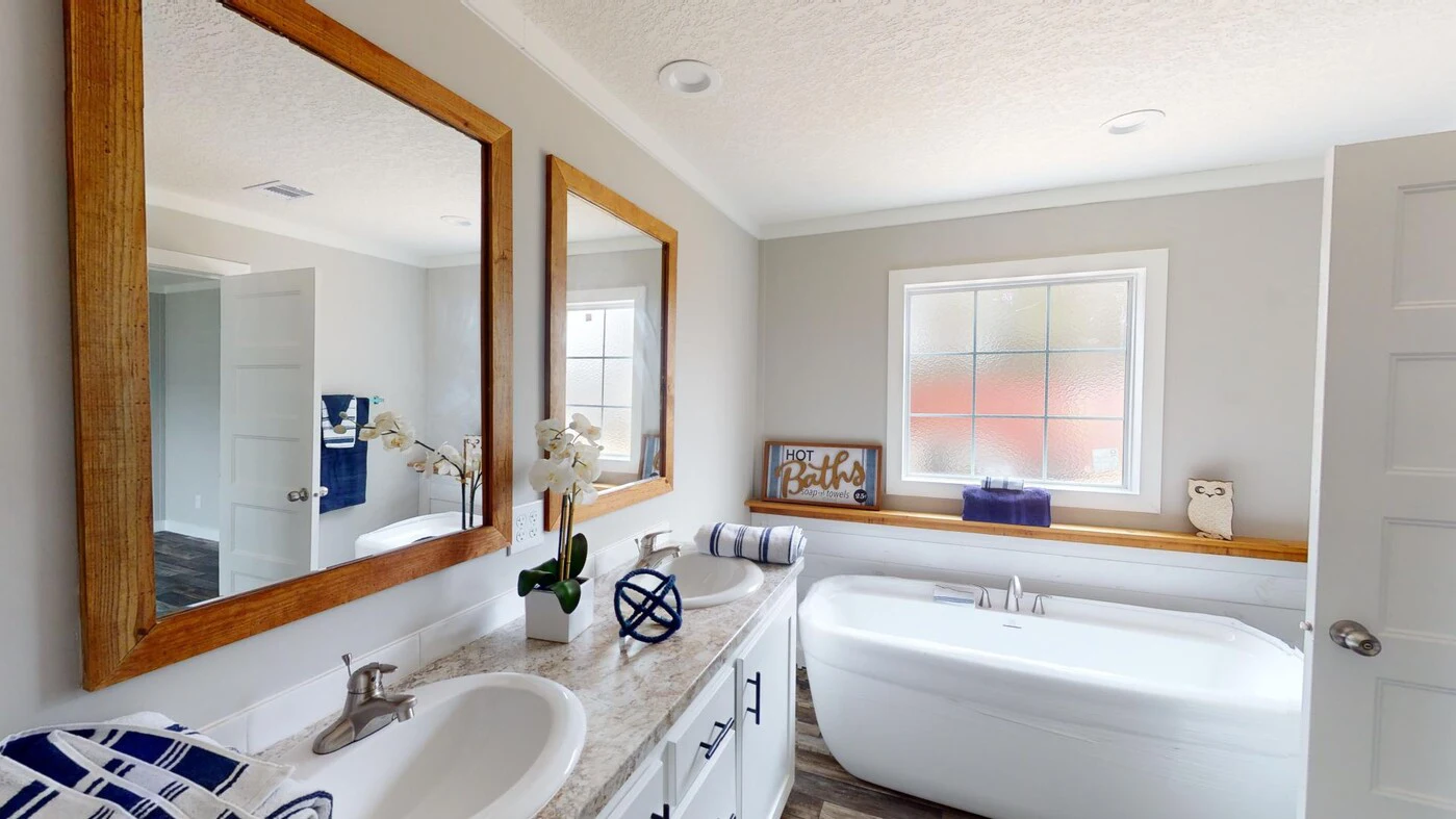 Modern bathroom with double sinks, wood-framed mirrors, a freestanding bathtub under a window, and decorative items on the counter and window ledge.