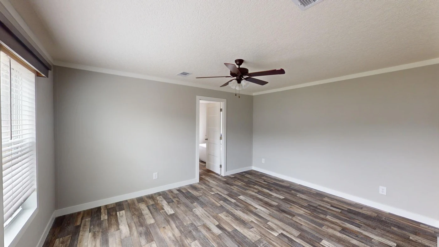 Empty room with light gray walls, a ceiling fan, large window with blinds, wood-look flooring, and a doorway leading to a white-tiled bathroom.