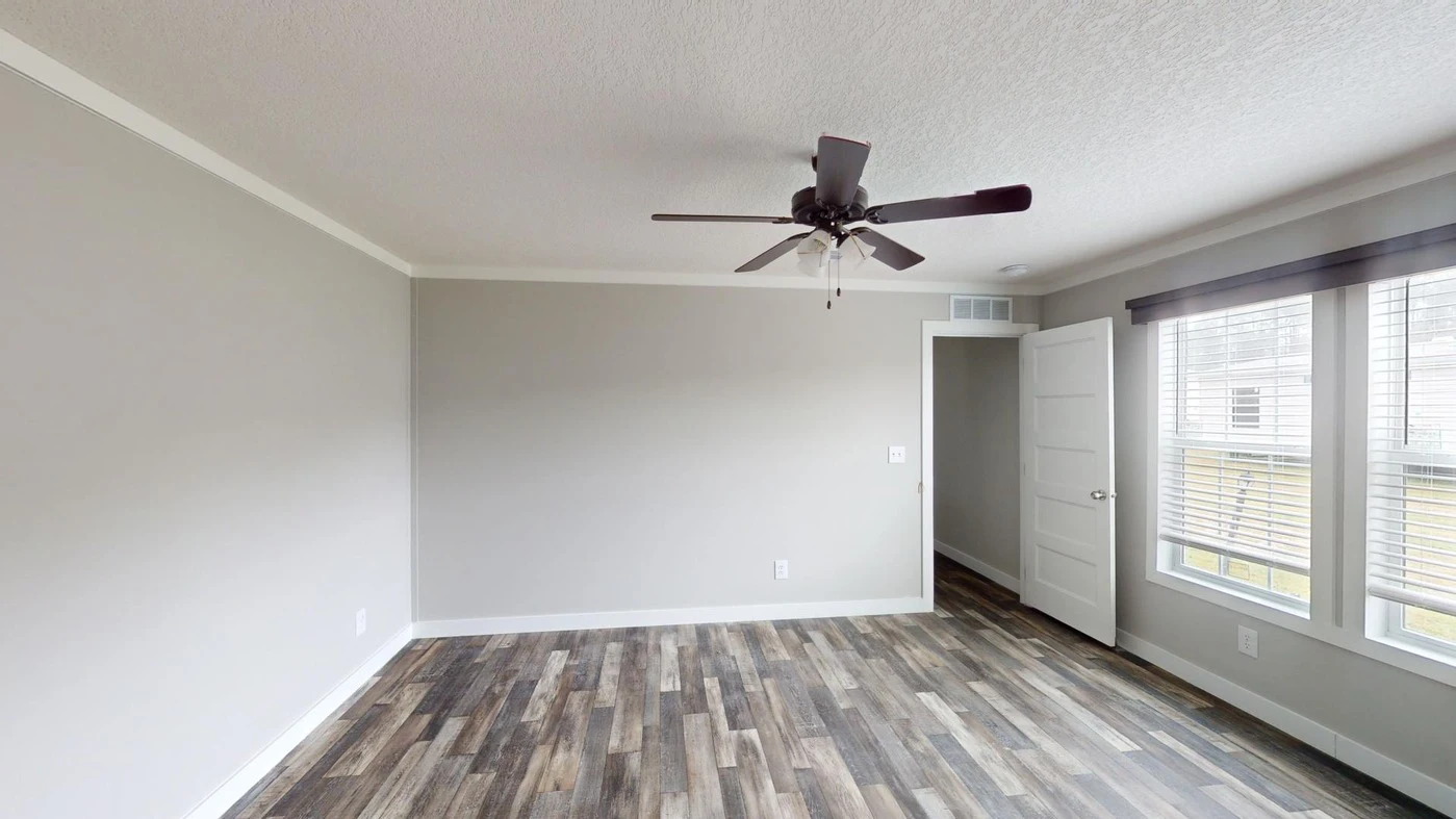 Empty room with light gray walls, wood-patterned flooring, a ceiling fan, three windows with blinds, and an open white door.