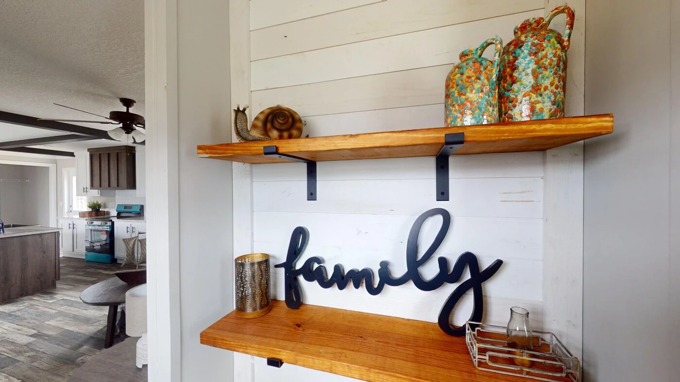Wooden shelves on a white shiplap wall hold decorative items, including vases, a snail ornament, a candle holder, and a "family" sign; kitchen visible in the background.
