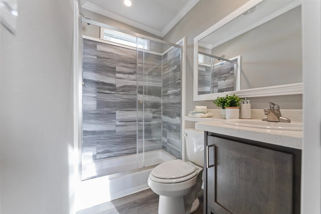 Modern bathroom with a glass-enclosed shower featuring gray tile, a toilet, a vanity with a sink, and a large mirror above the countertop.