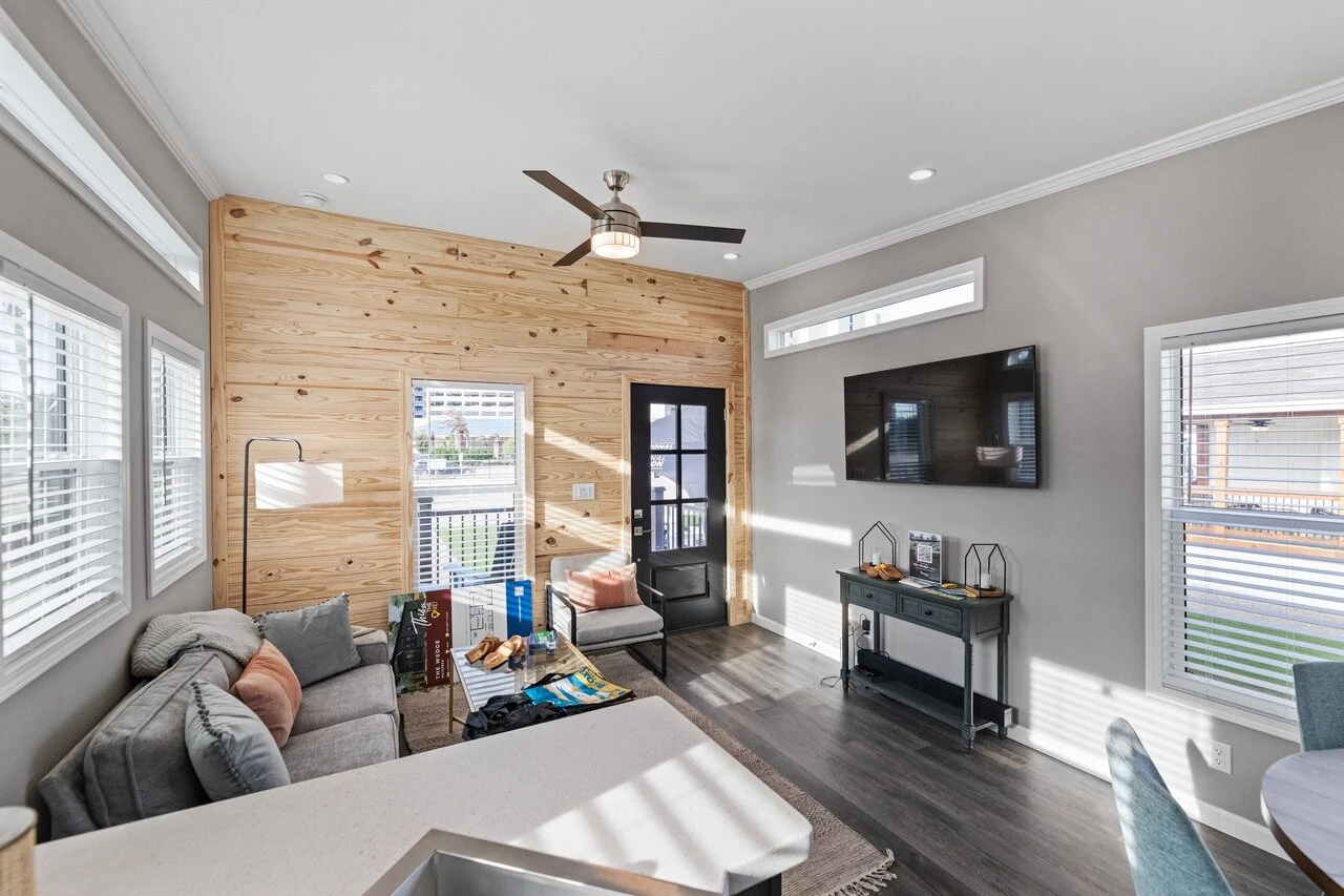 Modern living room with wood accent wall, gray sectional sofa, wall-mounted TV, ceiling fan, and large windows letting in natural light.