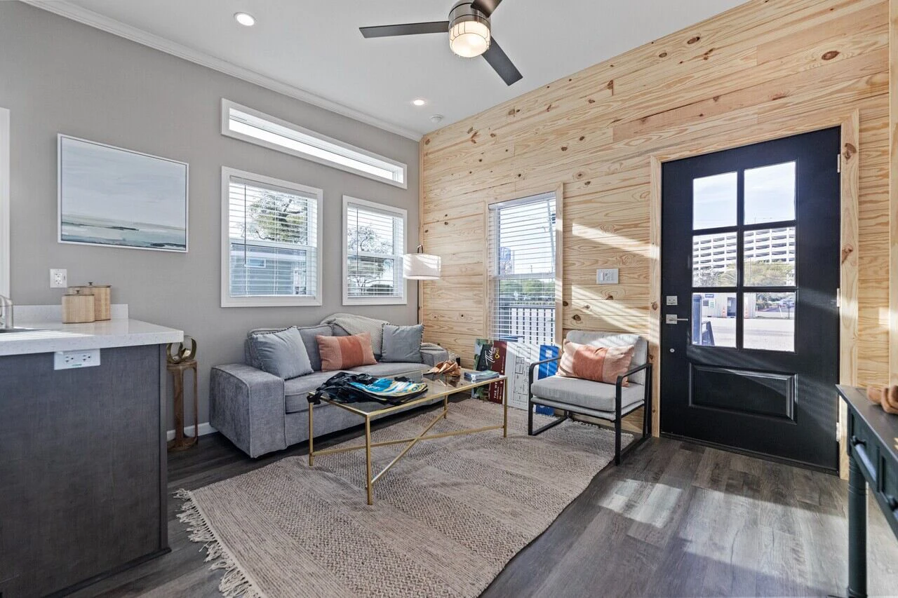 Modern living room with a gray sofa, armchair, and coffee table on a rug, featuring wood-paneled walls and a black door with windows letting in natural light.