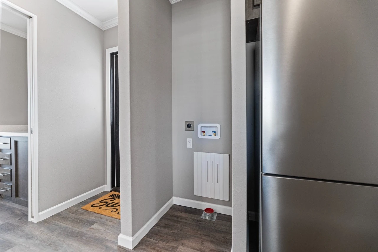 A laundry area with water hookups, an electrical outlet, and a vent on a gray wall, next to a refrigerator and adjacent to a hallway with gray flooring.